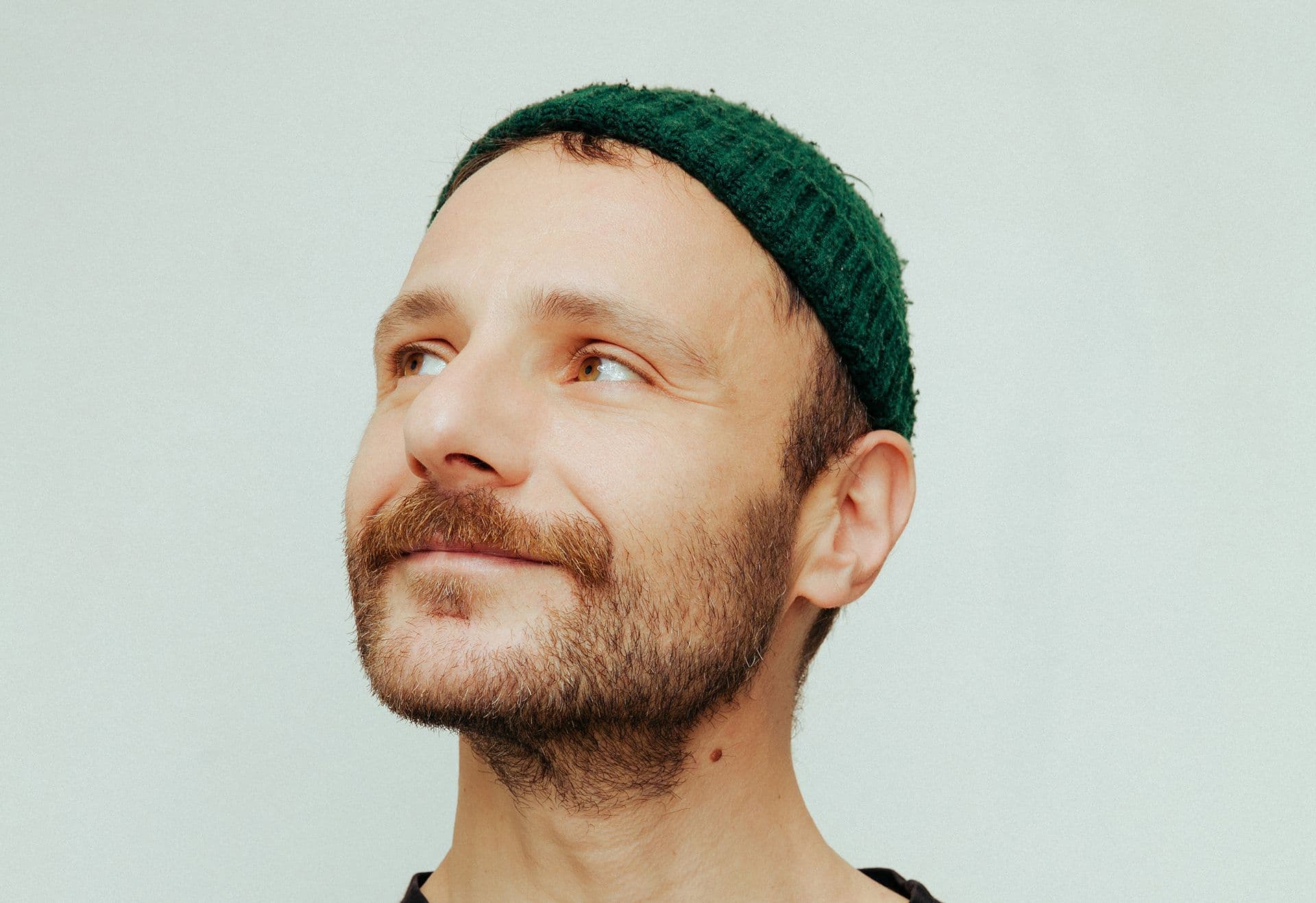 Ένας άνδρας με γένια και πράσινο σκουφάκι κοιτάζει ψηλά σε λευκό φόντο.