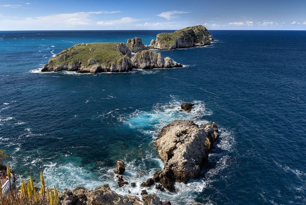 Felsige Inseln, umgeben von tiefblauem Ozean unter klarem Himmel, mit Wellen, die im Vordergrund gegen die Küste schlagen.