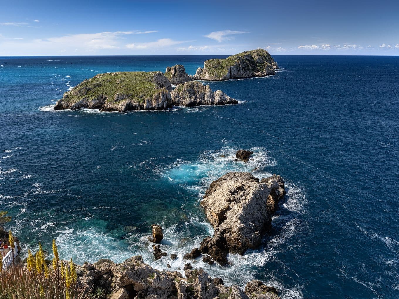Felsige Inseln, umgeben von tiefblauem Ozean unter klarem Himmel, mit Wellen, die im Vordergrund gegen die Küste schlagen.