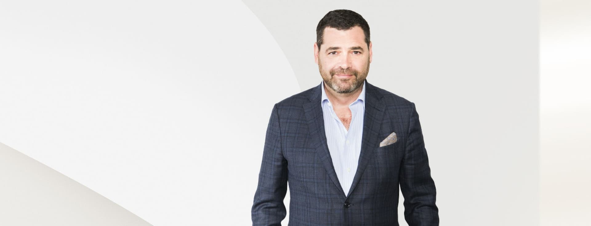 A man in a blue suit and light blue shirt stands against a white background.