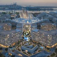 Aerial view of a futuristic cityscape at dusk with illuminated, curvy modern buildings, lush greenery, and waterfront in the background.