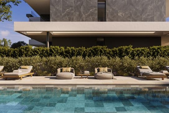 Sunlit turquoise pool with cushioned loungers and round chairs on a tiled deck beside a modern house.