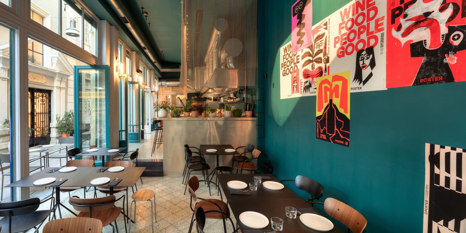 Εσωτερικό εστιατορίου με τοίχους τιρκουάζ, μαύρα τραπέζια και ξύλινες καρέκλες. Αφίσες κοσμούν τους τοίχους και φυτά διακοσμούν την κουζίνα. Φυσικό φως εισέρχεται άπλετα.