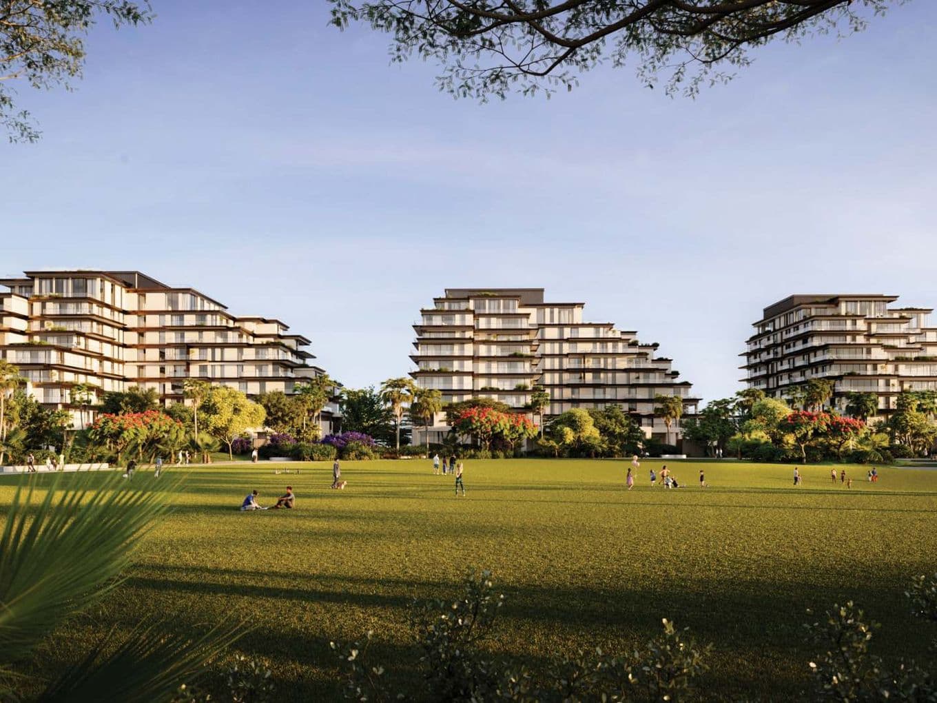 Modern, tiered buildings surrounded by lush greenery and an open lawn with people relaxing under a clear blue sky.