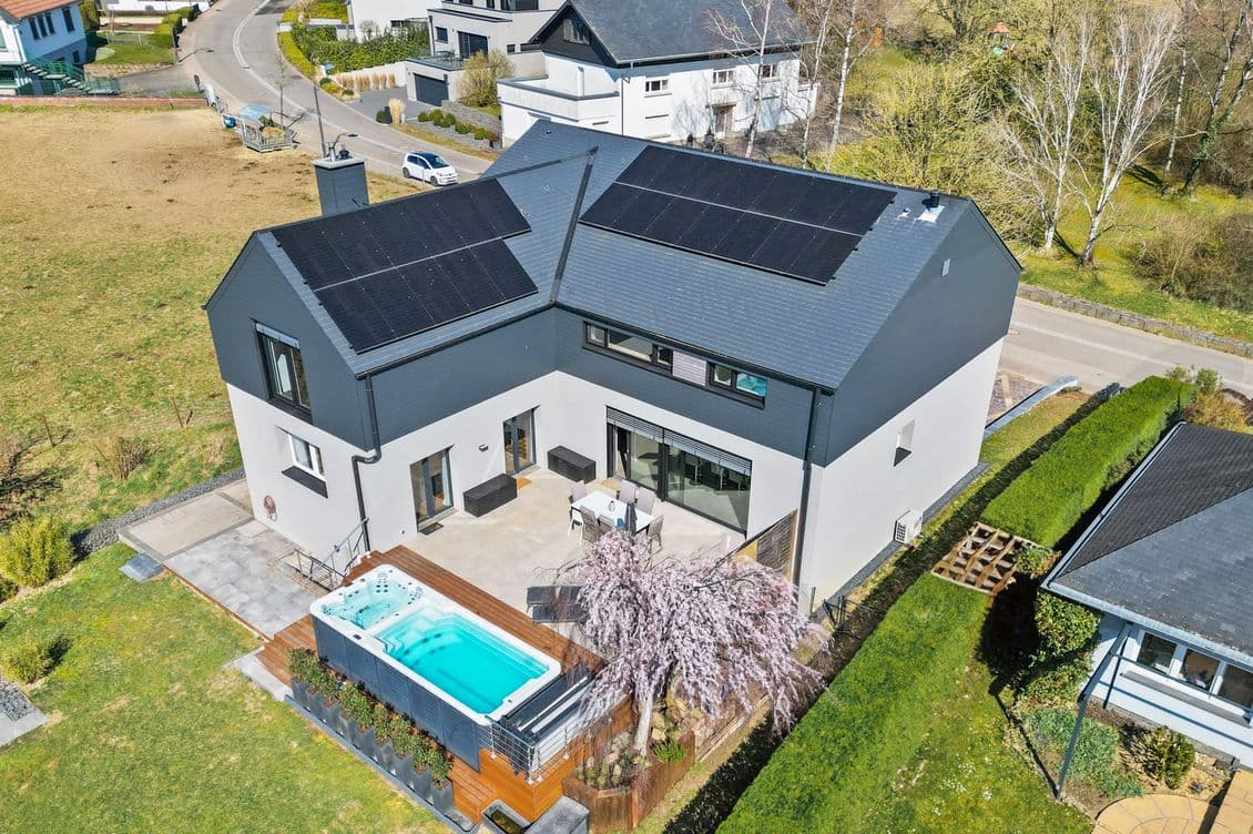 Aerial view of a modern white house with a dark roof and solar panels, a patio and pool, surrounded by a green yard on a quiet suburban street.