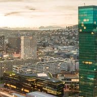 Luftaufnahme einer Stadtlandschaft bei Sonnenuntergang mit einem markanten Wolkenkratzer aus grünem Glas, umgeben von kleineren Gebäuden und Bahngleisen.