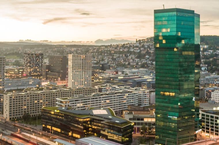 Luftaufnahme einer Stadtlandschaft bei Sonnenuntergang mit einem markanten Wolkenkratzer aus grünem Glas, umgeben von kleineren Gebäuden und Bahngleisen.