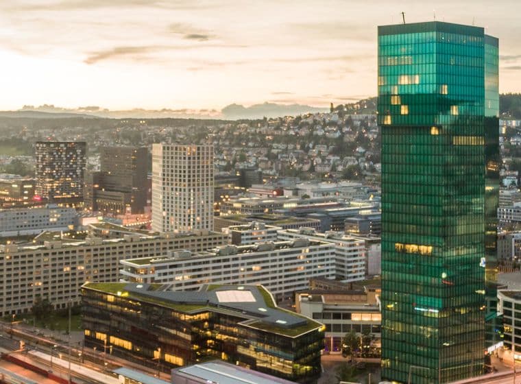 Luftaufnahme einer Stadtlandschaft bei Sonnenuntergang mit einem markanten Wolkenkratzer aus grünem Glas, umgeben von kleineren Gebäuden und Bahngleisen.