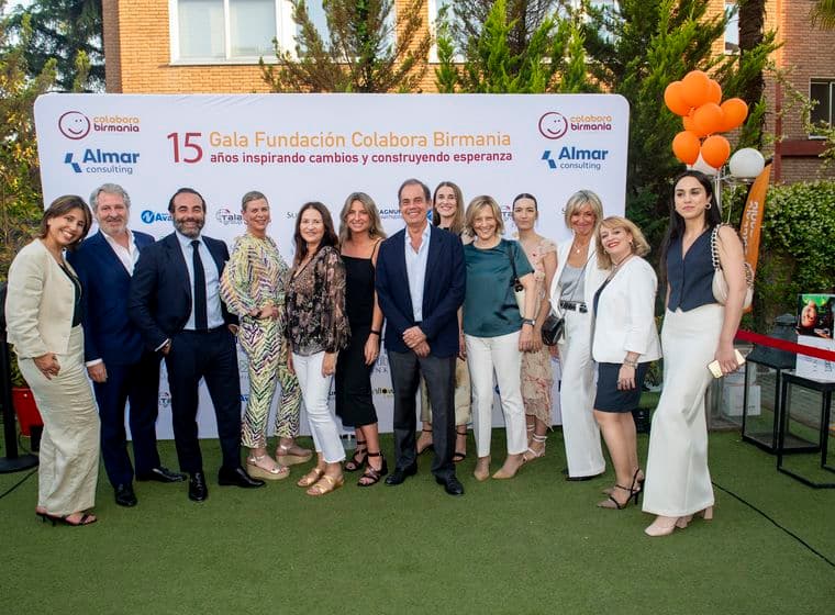 Un grup de 14 persones posen davant d'una pancarta de "Colabora Birmania" amb globus taronges. Vesteixen roba formal sobre una gespa verda.