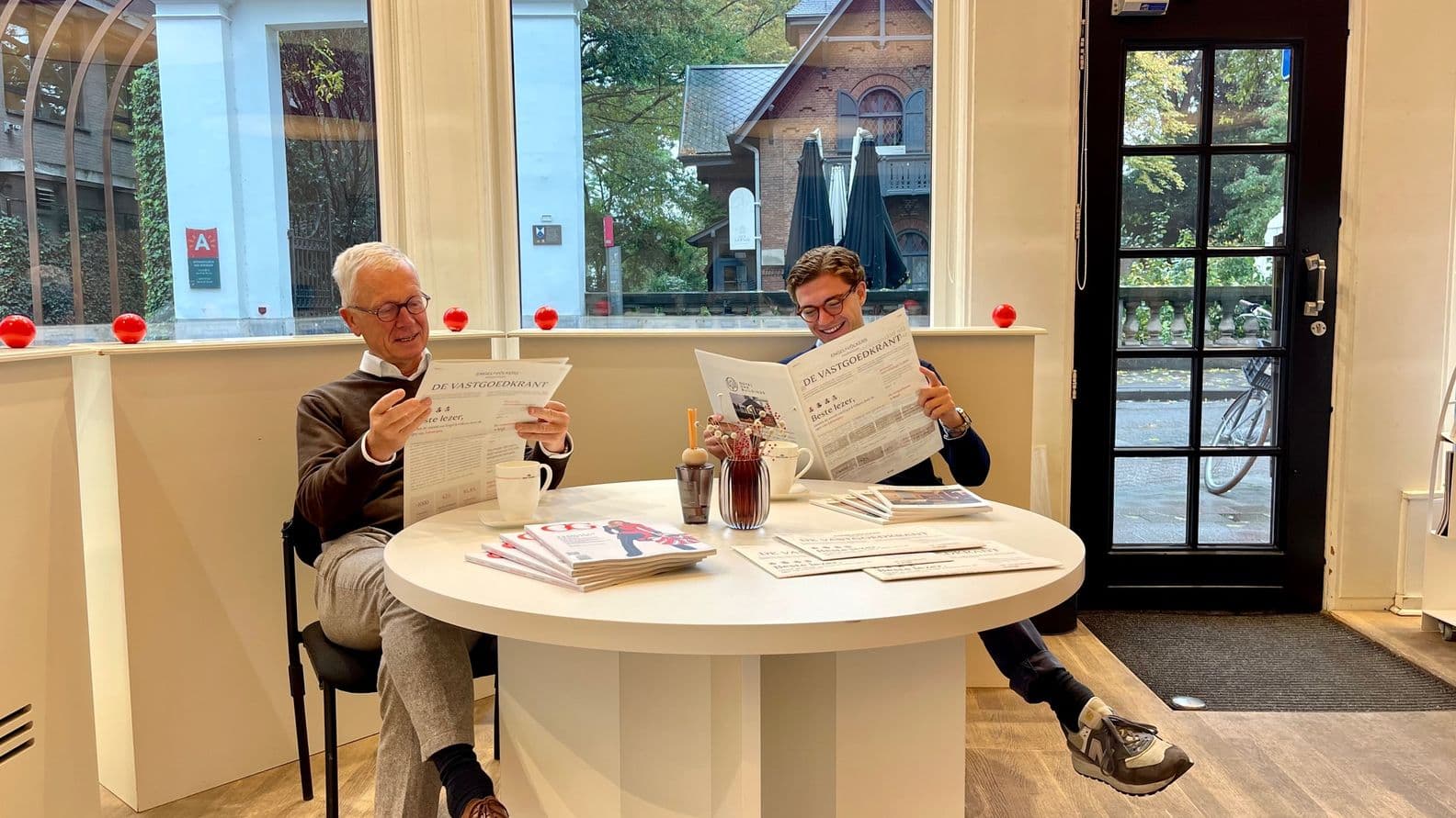 Two men reading newspapers at a white table in a real estate office with a view of a building.