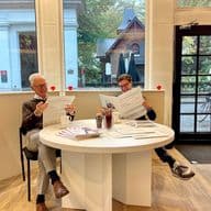 Twee mannen lezen kranten aan een witte tafel in een lichte kamer met grote ramen. Buiten zijn bomen en gebouwen zichtbaar.