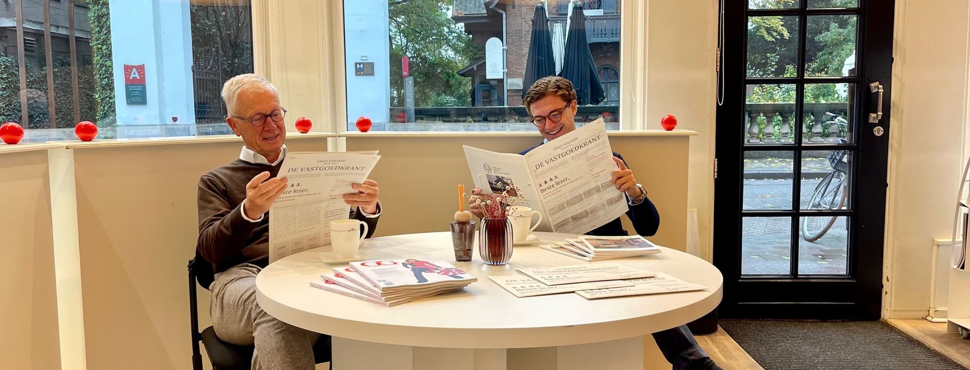 Twee mannen lezen kranten aan een witte tafel in een lichte kamer met grote ramen. Buiten zijn bomen en gebouwen zichtbaar.