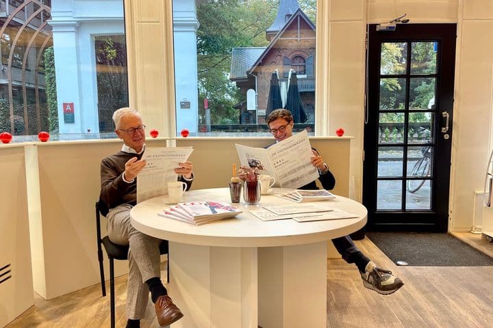 Twee mannen lezen kranten aan een witte tafel in een lichte kamer met grote ramen. Buiten zijn bomen en gebouwen zichtbaar.