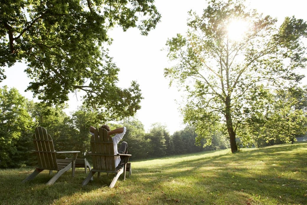 Two wooden chairs on a grassy field under trees, with sunlight streaming through branches, creating a peaceful, serene atmosphere.