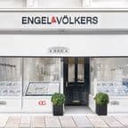 Frontansicht eines Engel & Völkers Immobilienbüros mit der Hausnummer 191, mit minimalistischer weißer Fassade, zentraler schwarzer Eingangstür, die von zwei Schaufenstern mit Immobilienangeboten flankiert wird, schwarzen Pflanzkübeln mit Buchsbaumkugeln und dem prominent über dem Eingang platzierten Firmenlogo sowie einer klassischen Villa-Illustration auf der Markise.