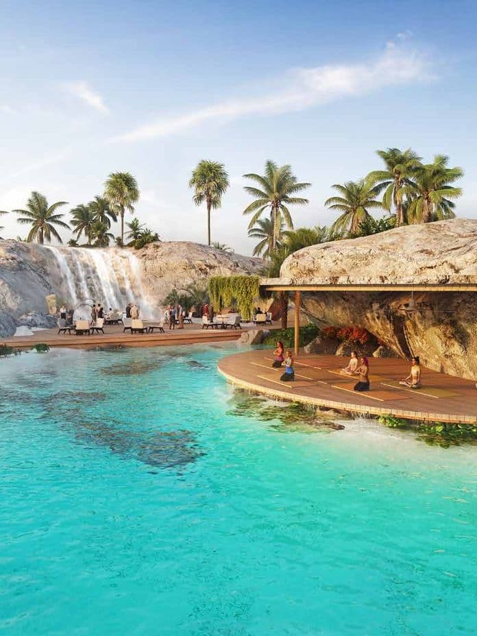 Tropical paradise with a waterfall, palm trees, clear blue water, and people relaxing on the beach and doing yoga on a deck.