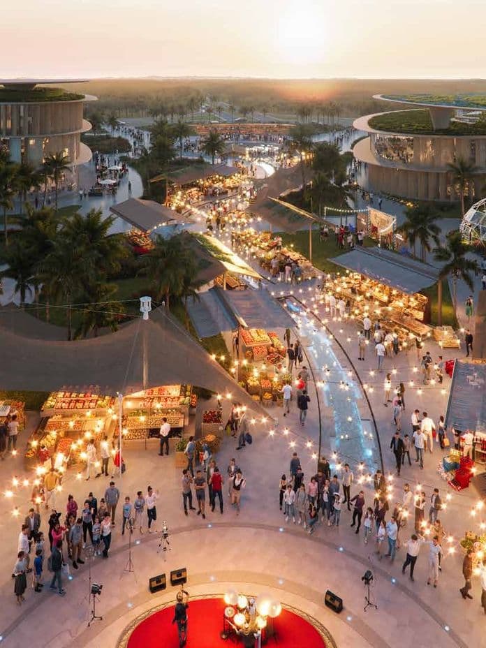 Aerial view of a vibrant outdoor market at sunset, with people, stalls, string lights, palm trees, and modern buildings in the background.