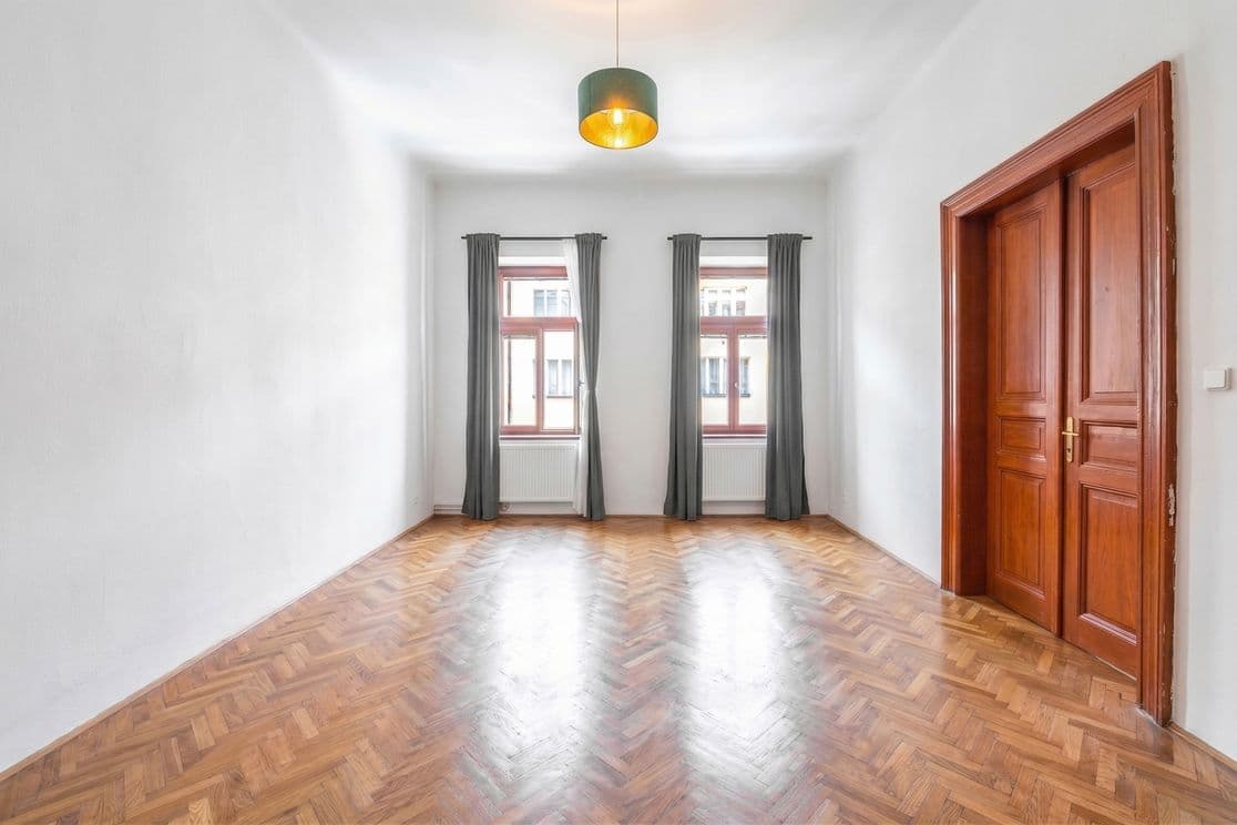 Empty room with white walls and herringbone-pattern parquet flooring. Two tall windows with dark grey curtains are positioned on the far wall, with radiators beneath them. A pendant light with a green shade and gold interior hangs from the centre of the ceiling. On the right side are original wooden double doors with decorative mouldings. The room feels bright, airy, and elegant, offering flexible use as a living space, bedroom, or home office.
