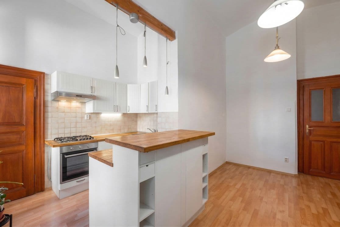 Spacious room with white walls, high ceilings, and light wood-effect flooring. On the left side is a kitchenette with white cabinetry, a gas cooktop, oven, and wooden worktops. In front of the kitchen stands a breakfast bar with integrated shelving and a raised wooden countertop. Modern pendant lights are suspended from a wooden beam above the kitchen area. The right side of the room remains open for living or dining use, while original wooden interior doors are visible along the wall. The interior feels airy, simple, and functional.