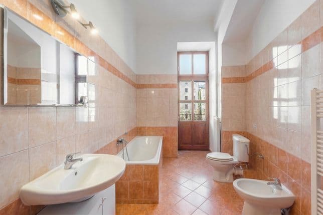 Bright bathroom with a high ceiling and ceramic wall tiles in beige and terracotta tones. On the left side are a washbasin with a mirrored cabinet and a built-in bathtub. On the right side are a toilet, bidet, and ladder-style towel radiator. At the far end of the room, glazed wooden doors lead to a balcony or gallery walkway, allowing natural daylight to enter the space. The floor is finished with matching ceramic tiles. The bathroom appears spacious, bright, and practical.