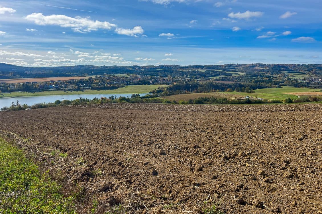 Ploughed field in the foreground, followed by open landscape with fields, a body of water, and scattered village buildings, with rolling hills in the background under a blue sky.