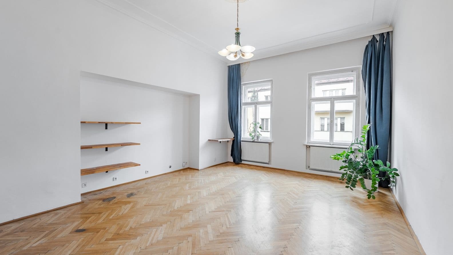 Empty room with white walls, high ceilings, and light herringbone parquet flooring. Two tall windows with dark blue curtains are positioned on the far wall, with radiators beneath the sills. On the left side of the room is a deep recessed niche fitted with three wooden wall-mounted shelves. A decorative pendant light hangs from a ceiling rose at the centre of the room. A potted plant stands near the right wall. The interior feels bright, airy, and elegant, with flexible potential for living, sleeping, or working purposes.