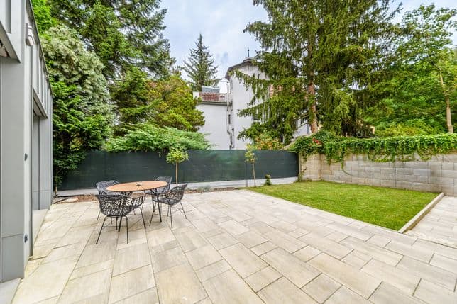 Přízemní terasa se stolem a čtyřmi židlemi, navazující travnatá plocha, kolem vzrostlé stromy a zídka porostlá popínavými rostlinami.