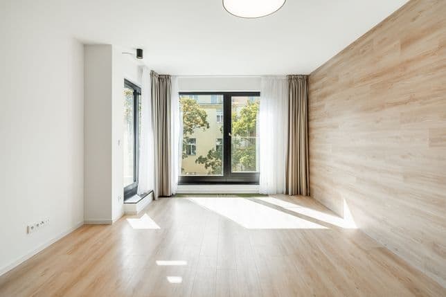 Prázdný pokoj s dřevěnou podlahou, světlým obkladem na jedné stěně, velkými okny s bílými a béžovými závěsy a výhledem na stromy.