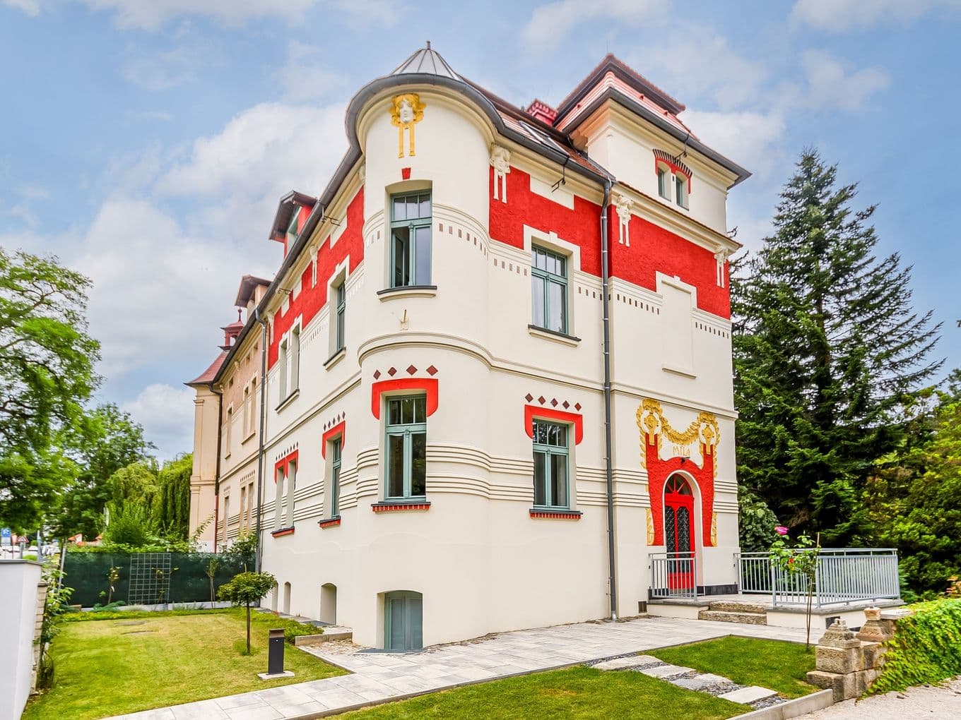 Třípodlažní secesní vila s červenobílou fasádou, zdobená ornamenty a nápisem „Vila“, obklopená trávníkem a stromy.