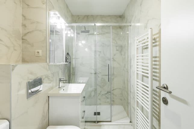 Koupelna s mramorovým obkladem, sprchovým koutem s prosklenými dveřmi, bílým umyvadlem se skříňkou, zrcadlem a bílým topným žebříkem.
