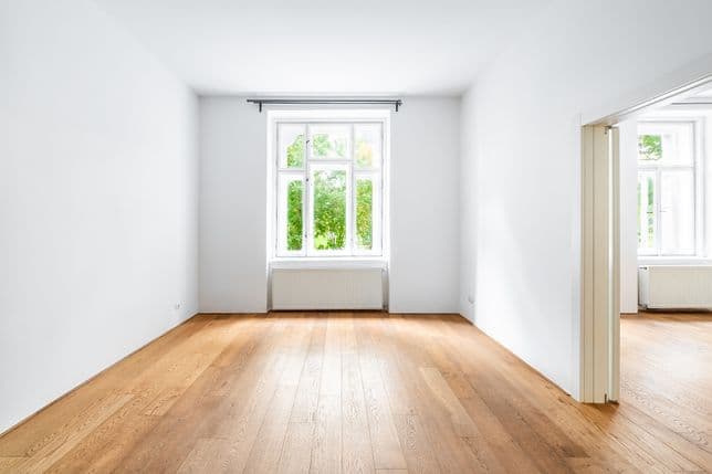 Prázdný pokoj s dřevěnou podlahou, bílými stěnami, velkým oknem s výhledem do zeleně a průchodem do další místnosti se stejným oknem.