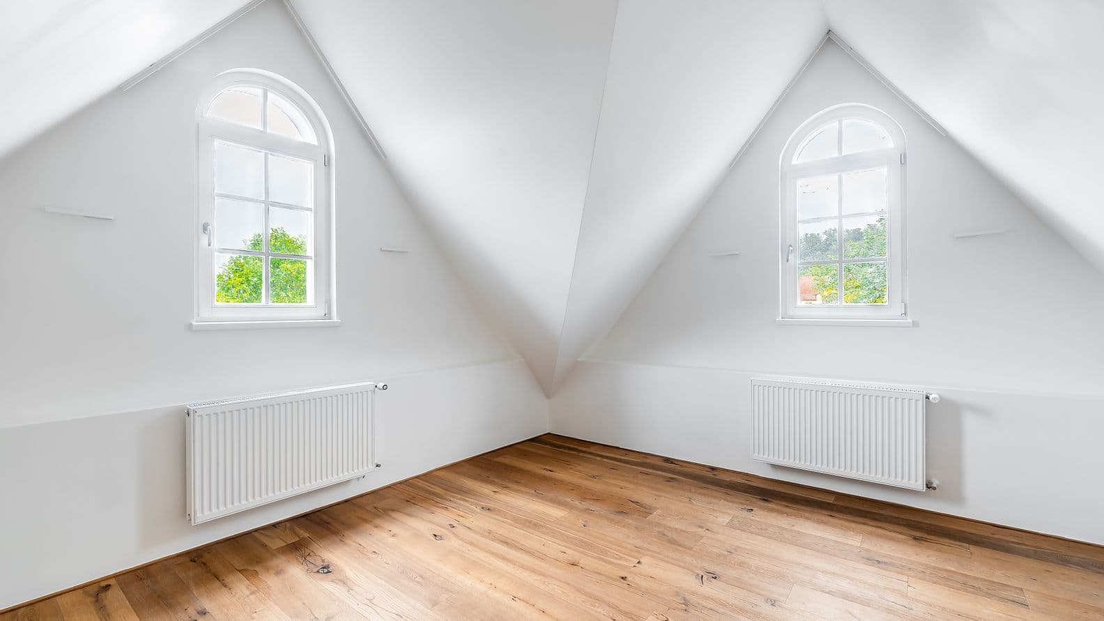 Prázdný podkrovní pokoj s dřevěnou podlahou, bílými stěnami a dvěma obloukovými okny, ze kterých je výhled na stromy.