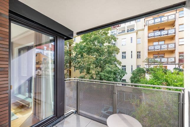 Balkon s kovovým zábradlím a malým stolkem, navazující na obývací pokoj, s výhledem na stromy a okolní domy se žlutými fasádami.