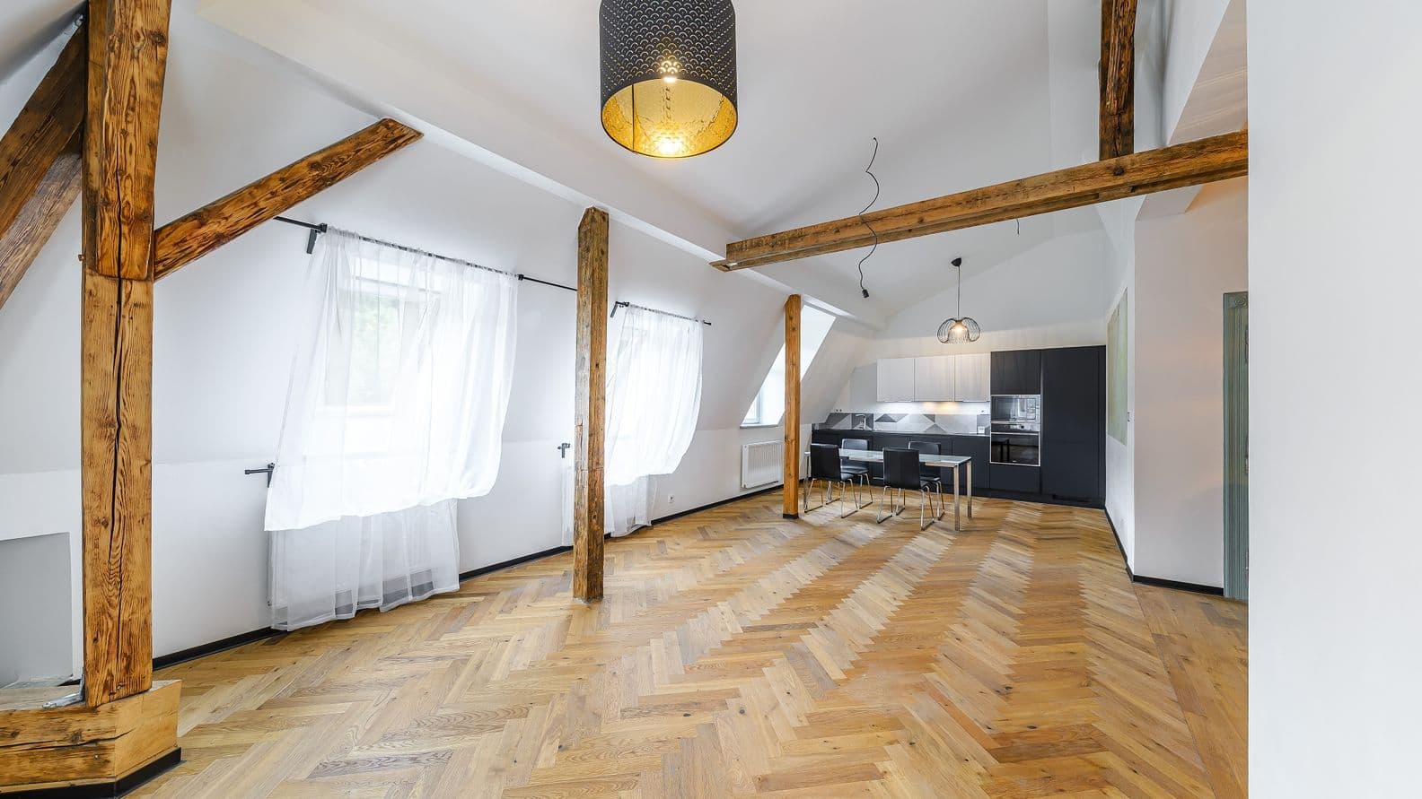 Podkrovní místnost s dřevěnou podlahou, odhalenými trámy, několika okny se záclonami a kuchyňským koutem s jídelním stolem.