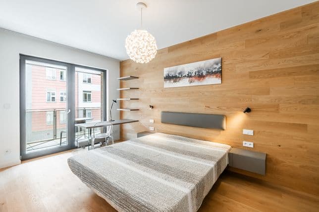 Ložnice s dřevěnou stěnou, dvojlůžkem s šedým čelem, psacím stolem a policemi u okna, odkud je výhled na sousední budovy.
