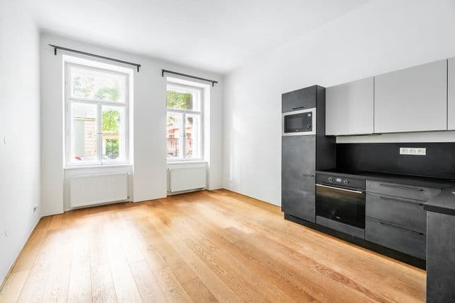 Místnost s kuchyňskou linkou v černé a šedé barvě, dvěma okny směrem do ulice a světlou dřevěnou podlahou.