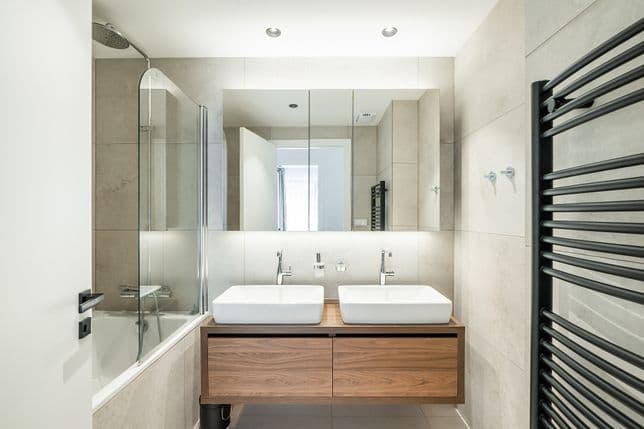 Koupelna s velkým zrcadlem, dvěma umyvadly na dřevěné skříňce, vanou se skleněnou zástěnou a černým topným žebříkem.