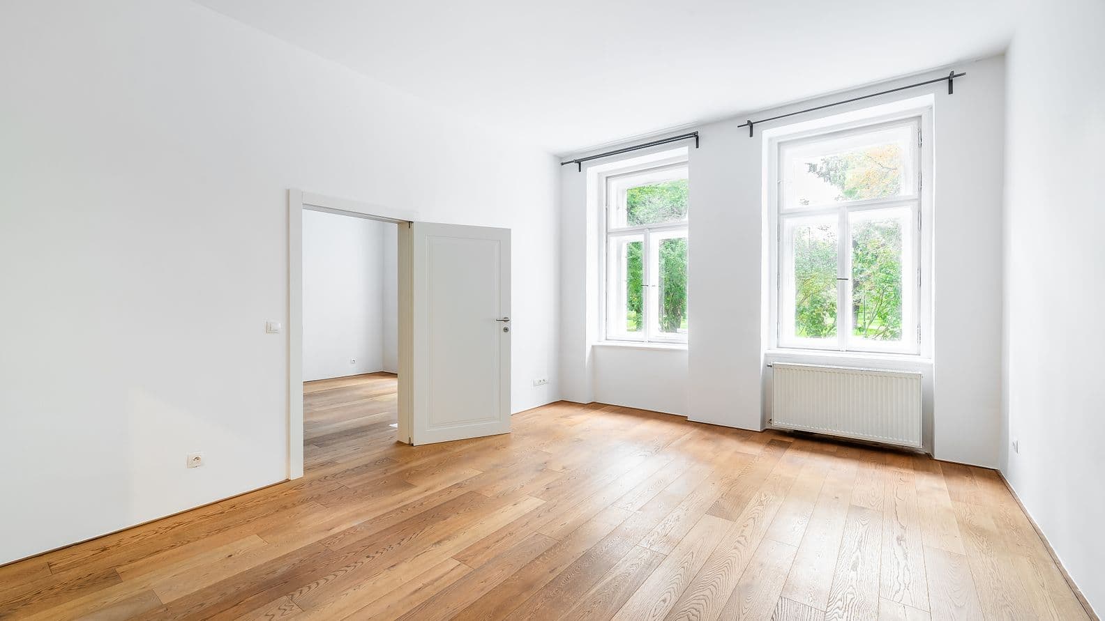 Světlý pokoj s dřevěnou podlahou, dvěma okny do parku, bílými stěnami a otevřenými dveřmi vedoucími do další místnosti se stejnou podlahou.