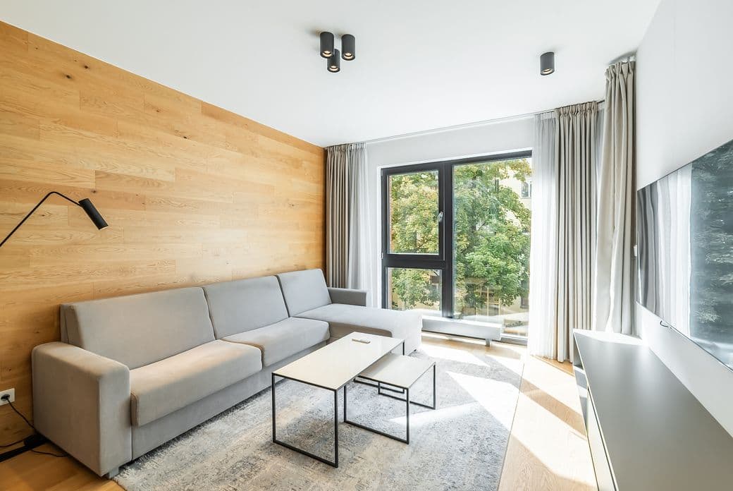 Living room with a grey corner sofa, wooden accent wall, two coffee tables, and a large window overlooking trees.