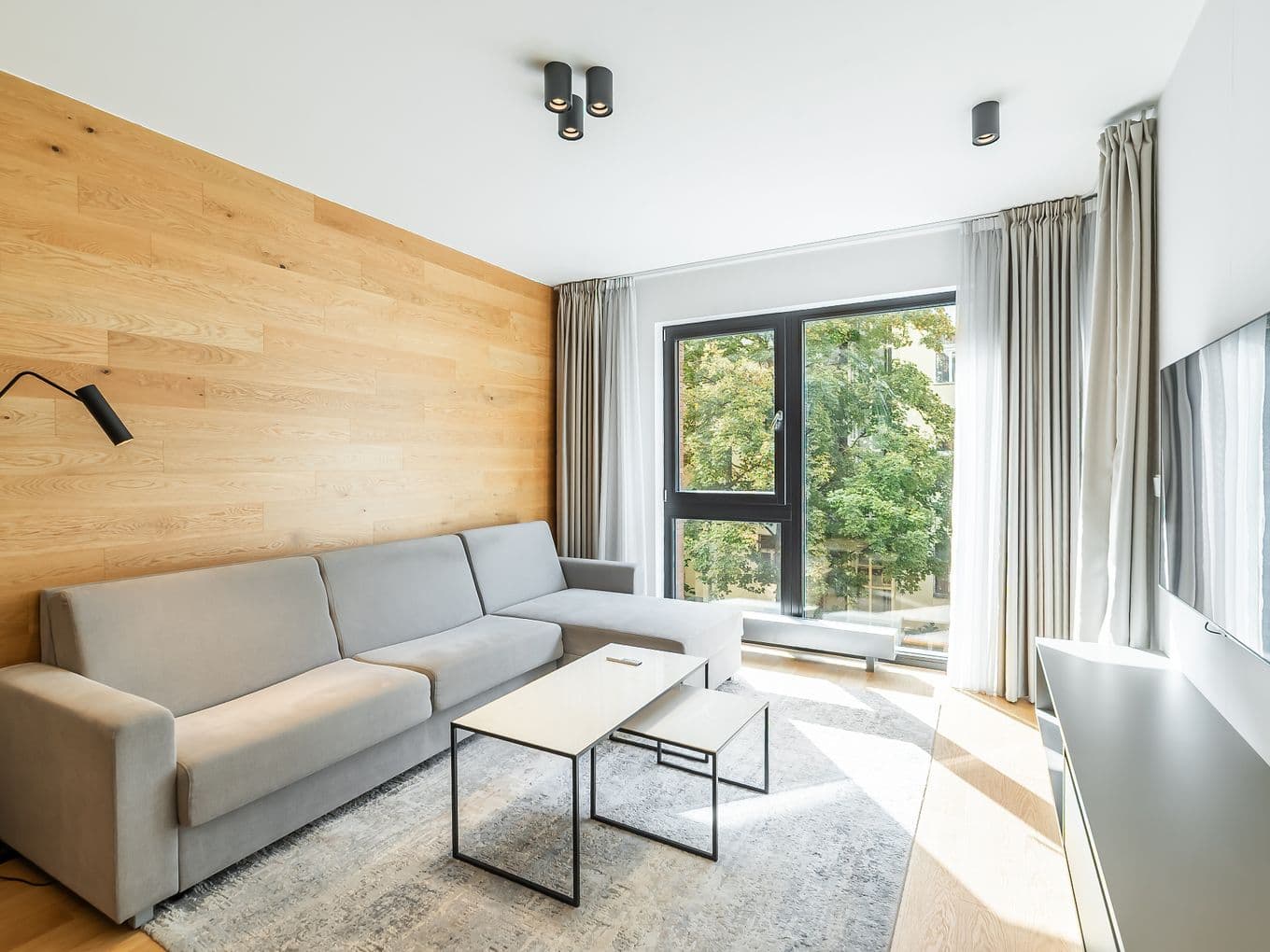 Obývací pokoj s šedou rohovou pohovkou, dřevěným obkladem na stěně, dvěma konferenčními stolky a velkým oknem s výhledem na stromy.