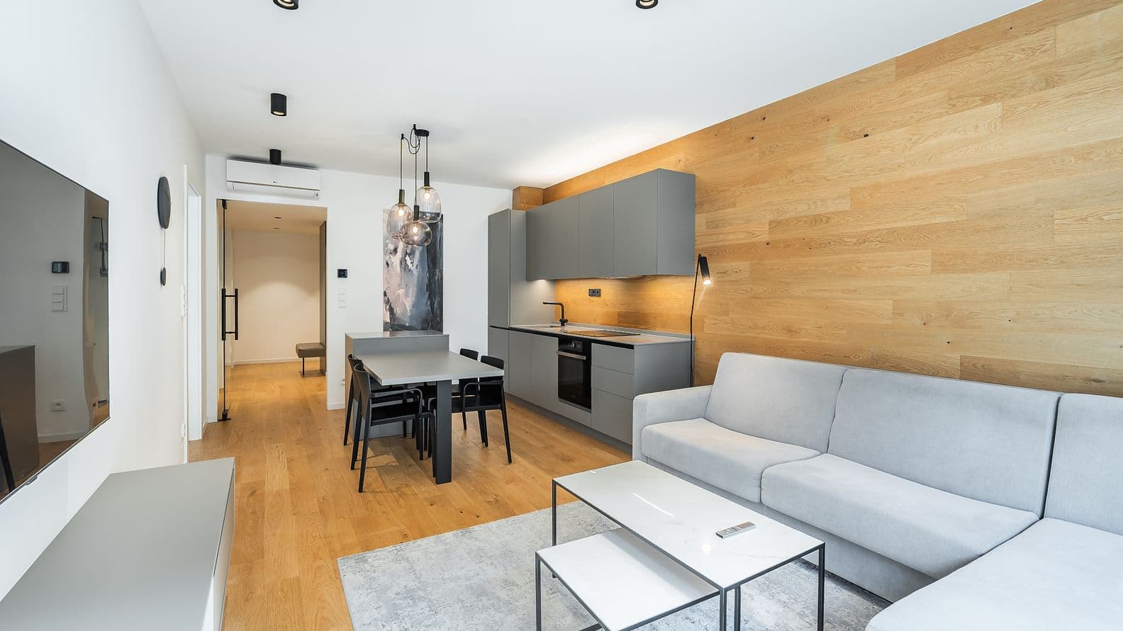 Světlý obývací pokoj s šedou rohovou pohovkou, dřevěnou podlahou, kuchyňskou linkou v šedé barvě a jídelním stolem se čtyřmi černými židlemi, osvětlený závěsnými lampami.