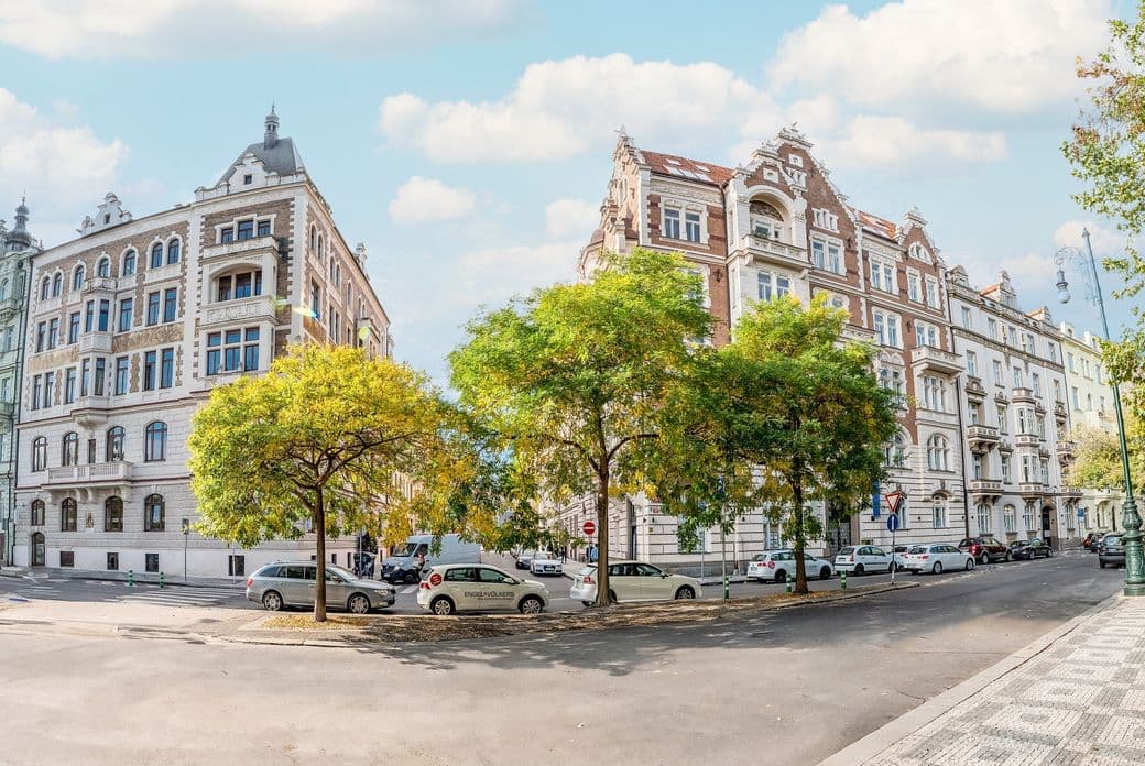 Historic Prague buildings with ornate facades line a street, shaded by leafy trees, under a partly cloudy sky.