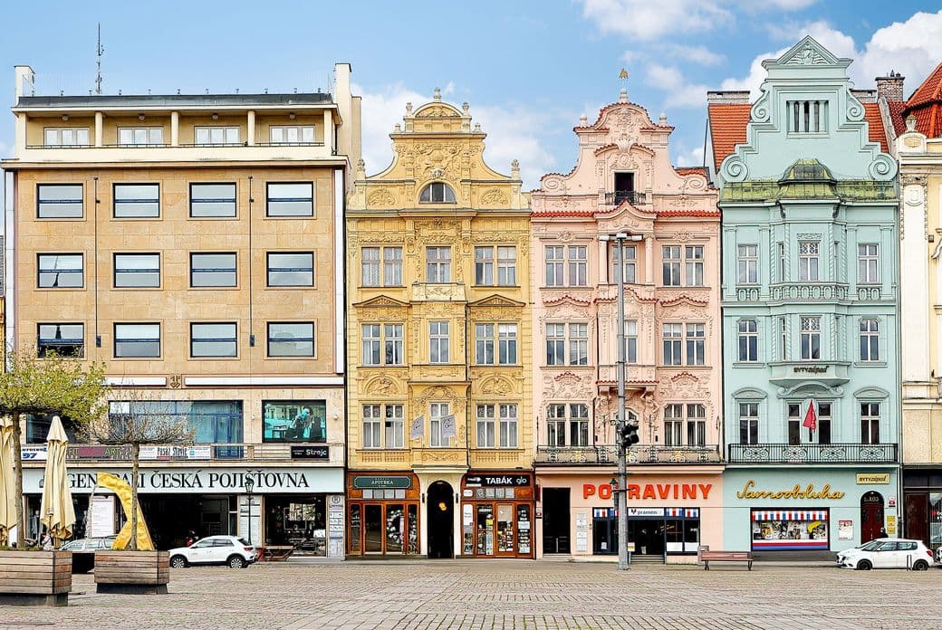 A row of buildings on Republic Square in Plzeň with colorful pastel facades, combining historical and modernist architecture. The ground floors feature shops and local businesses.