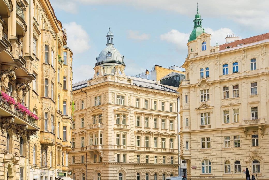 A street in central Prague lined with ornate historic buildings in neoclassical and Art Nouveau styles. In the foreground, a building with a dome and another with a green turret, both typical of Prague architecture.