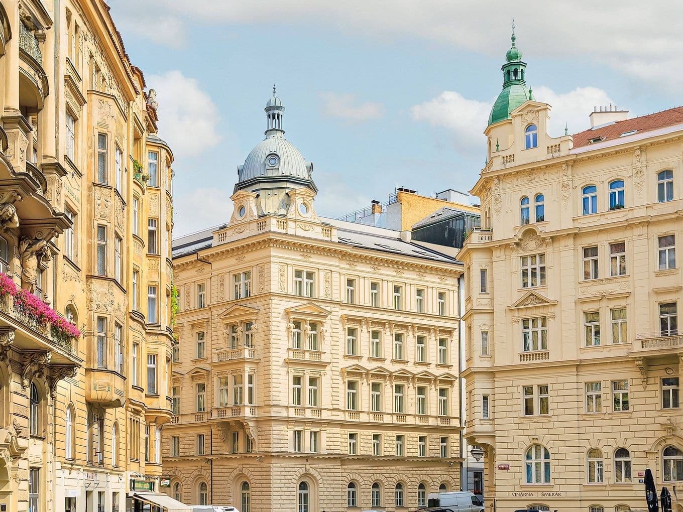 Ulice v centru Prahy lemovaná zdobenými historickými budovami v neoklasickém a secesním stylu. V popředí se nachází budova s kupolí a další se zelenou věžičkou, obě typické pro pražskou architekturu.