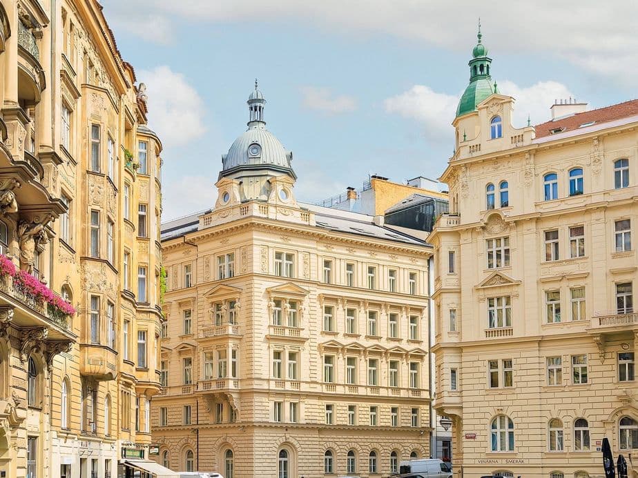 A street in central Prague lined with ornate historic buildings in neoclassical and Art Nouveau styles. In the foreground, a building with a dome and another with a green turret, both typical of Prague architecture.
