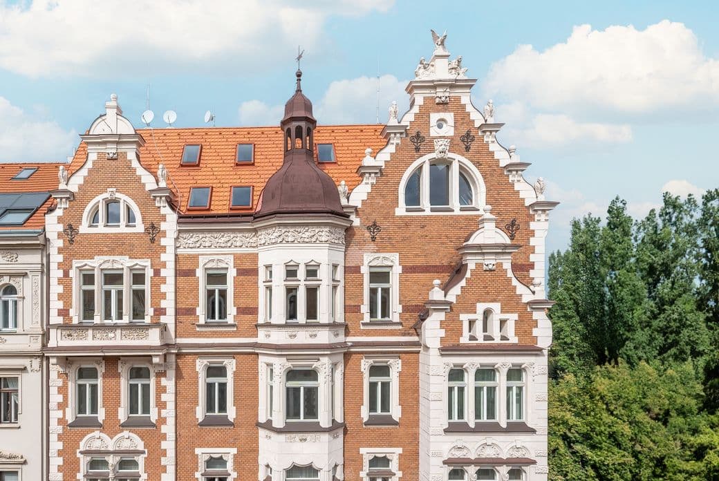 Detailed view of a historic apartment building with a red brick facade, white stucco elements, and an ornate turret. A typical example of Art Nouveau architecture in Prague, with a mansard roof and rich decoration.