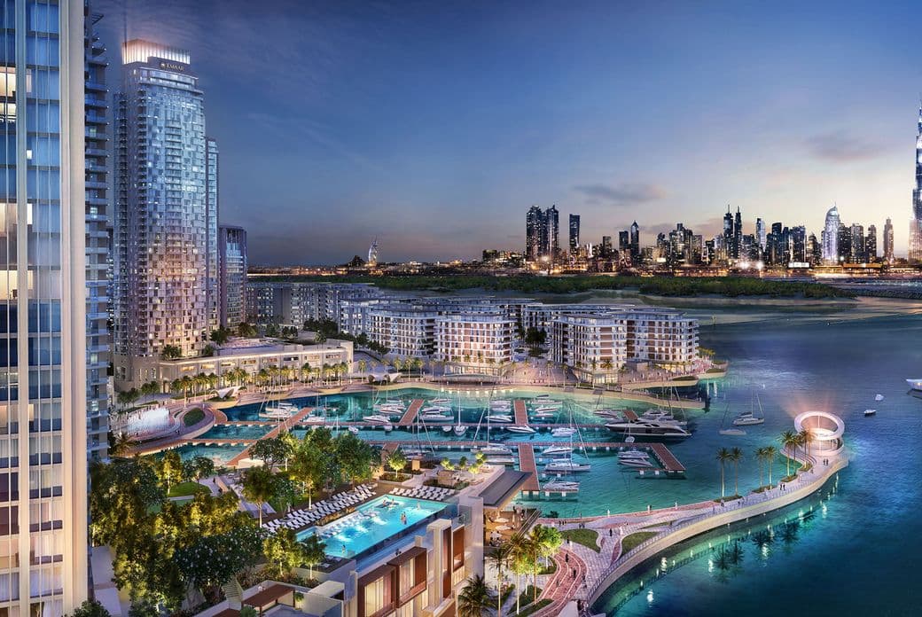 City skyline at dusk with illuminated skyscrapers, a marina with boats, and modern waterfront buildings surrounded by water.