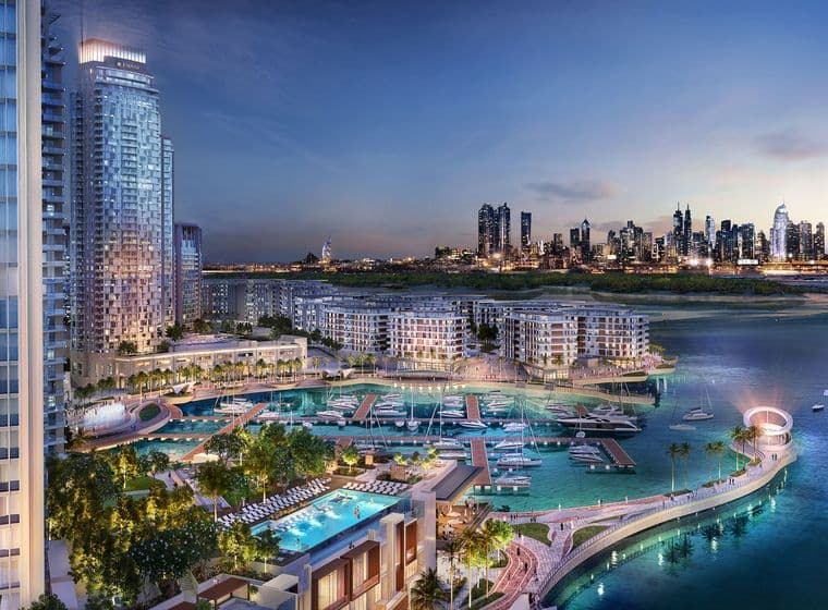 City skyline at dusk with illuminated skyscrapers, a marina with boats, and modern waterfront buildings surrounded by water.