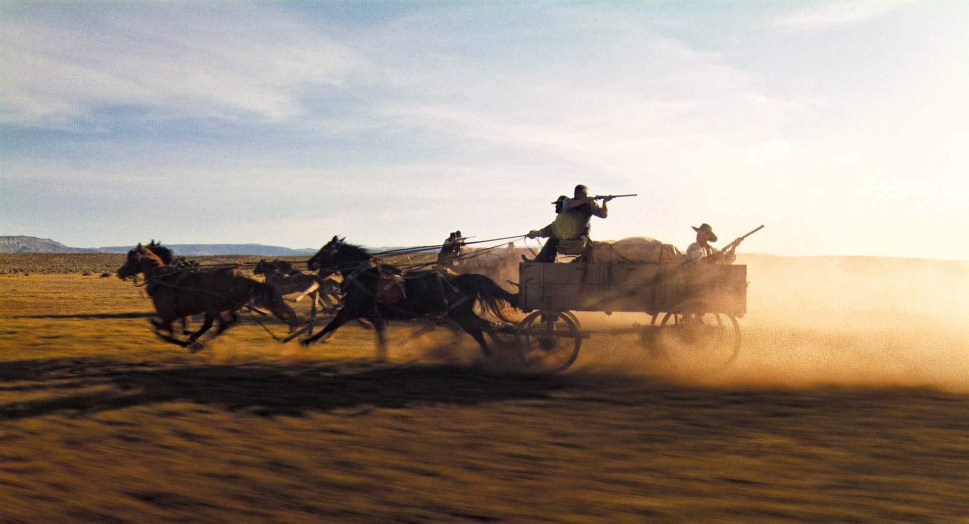 A horse-drawn wagon races across a dry, grassy plain, with armed men riding and shooting.